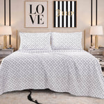 A neatly made bed with a white and gray geometric patterned quilt is flanked by two nightstands with lamps. The bed features a luxury bedding ensemble, complemented by the Microfiber Sheet Set With Extra Pillowcases for added comfort. Above the bed, two framed art pieces reading "LOVE" and "Juicy" adorn the wall.
