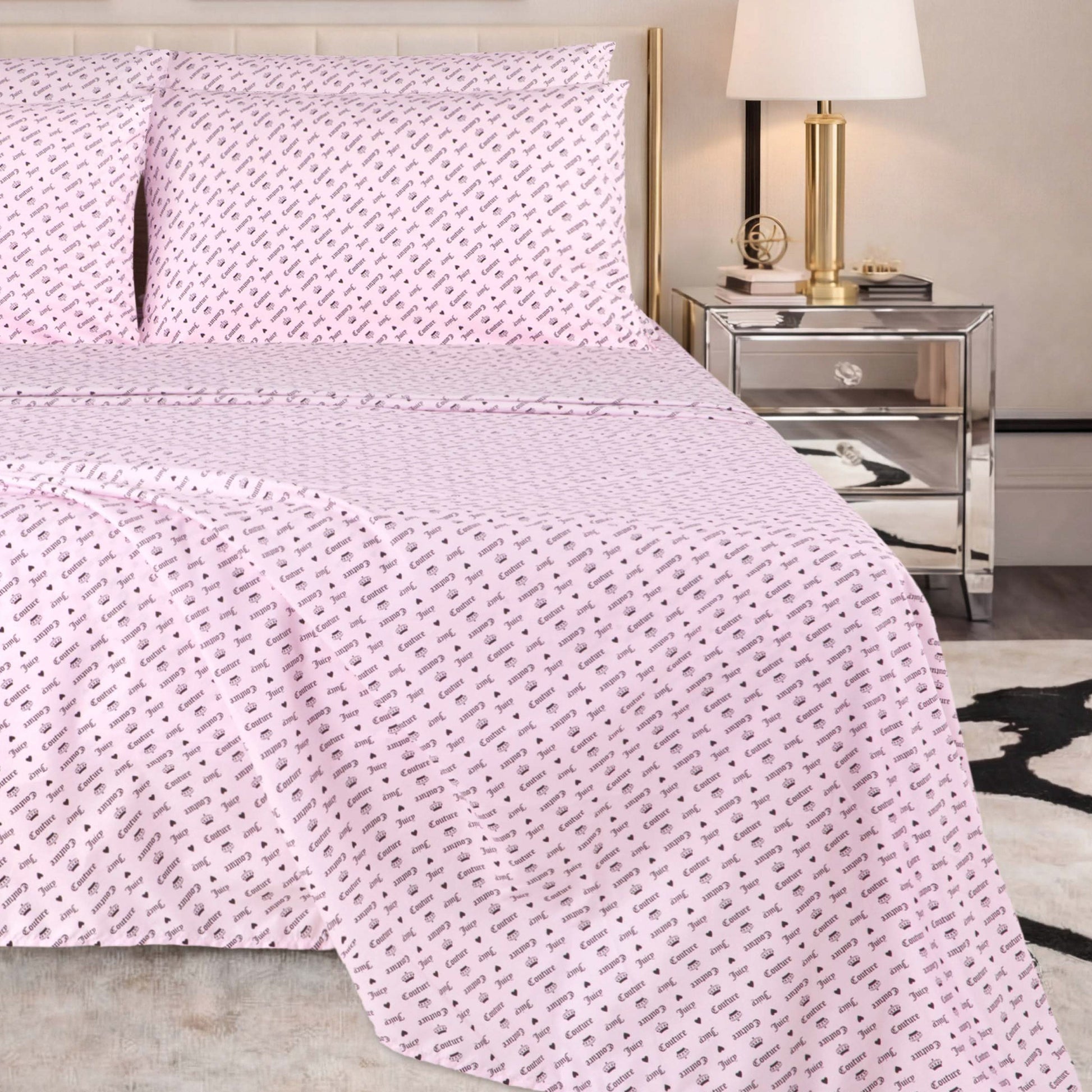 A neatly made bed with a pale pink Microfiber Sheet Set With Extra Pillowcases, featuring a small repeating black pattern, is situated next to a mirrored nightstand with a gold lamp and books in a modern bedroom setting.