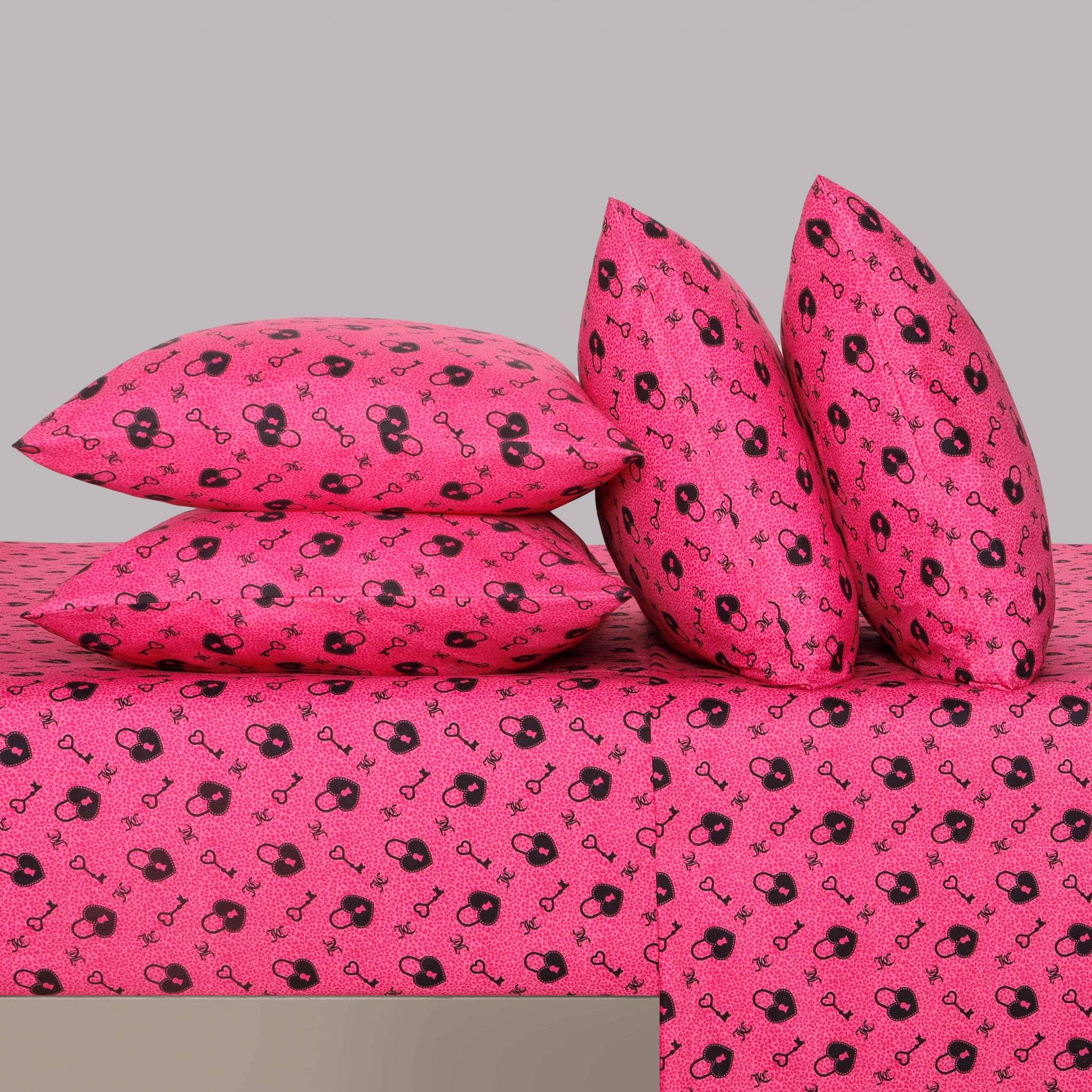 Four pillows—two stacked and two upright—all covered in vibrant pink fabric featuring a repeating pattern of black handcuffs and masks, are arranged on a matching Microfiber Sheet Set With Extra Pillowcases against a plain gray background.