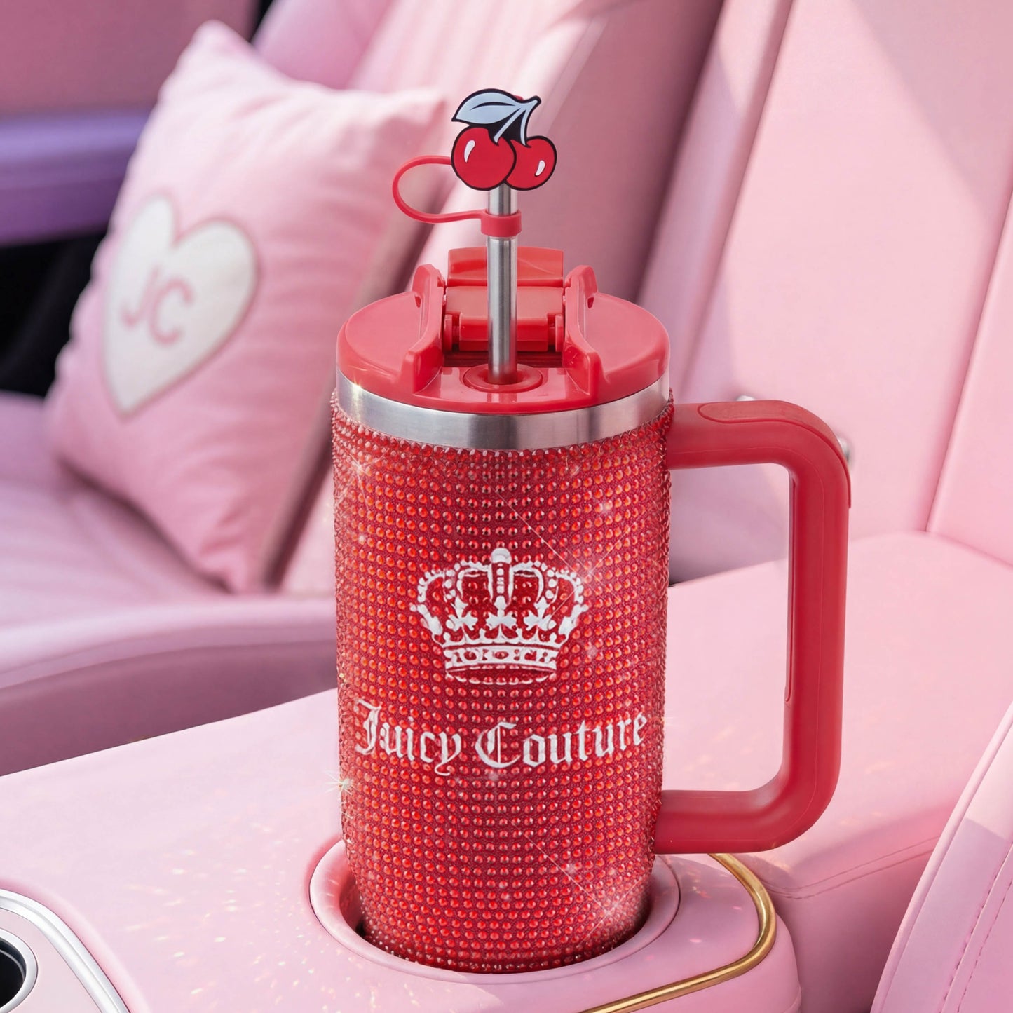 A red Diamond Bling Stainless Steel Tumbler with Handle and Straw, 30 oz, covered in rhinestones and featuring a cherry straw, sits in a pink car interior. A heart-shaped pillow with "JC" is visible in the background.