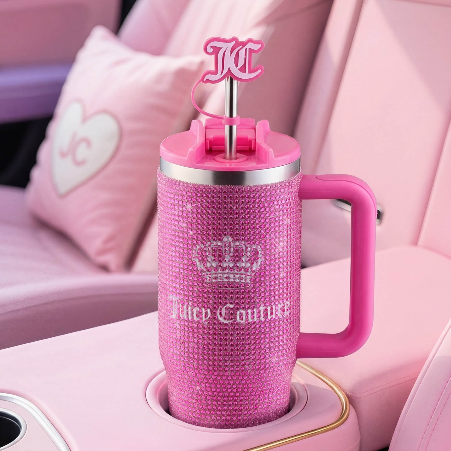 A Diamond Bling Stainless Steel Tumbler with Handle and Straw, 30 oz, in sparkling pink sits in a pink car interior. The tumbler features rhinestone accents, a crown logo, prominent “Juicy Couture” text, and a JC logo straw topper.