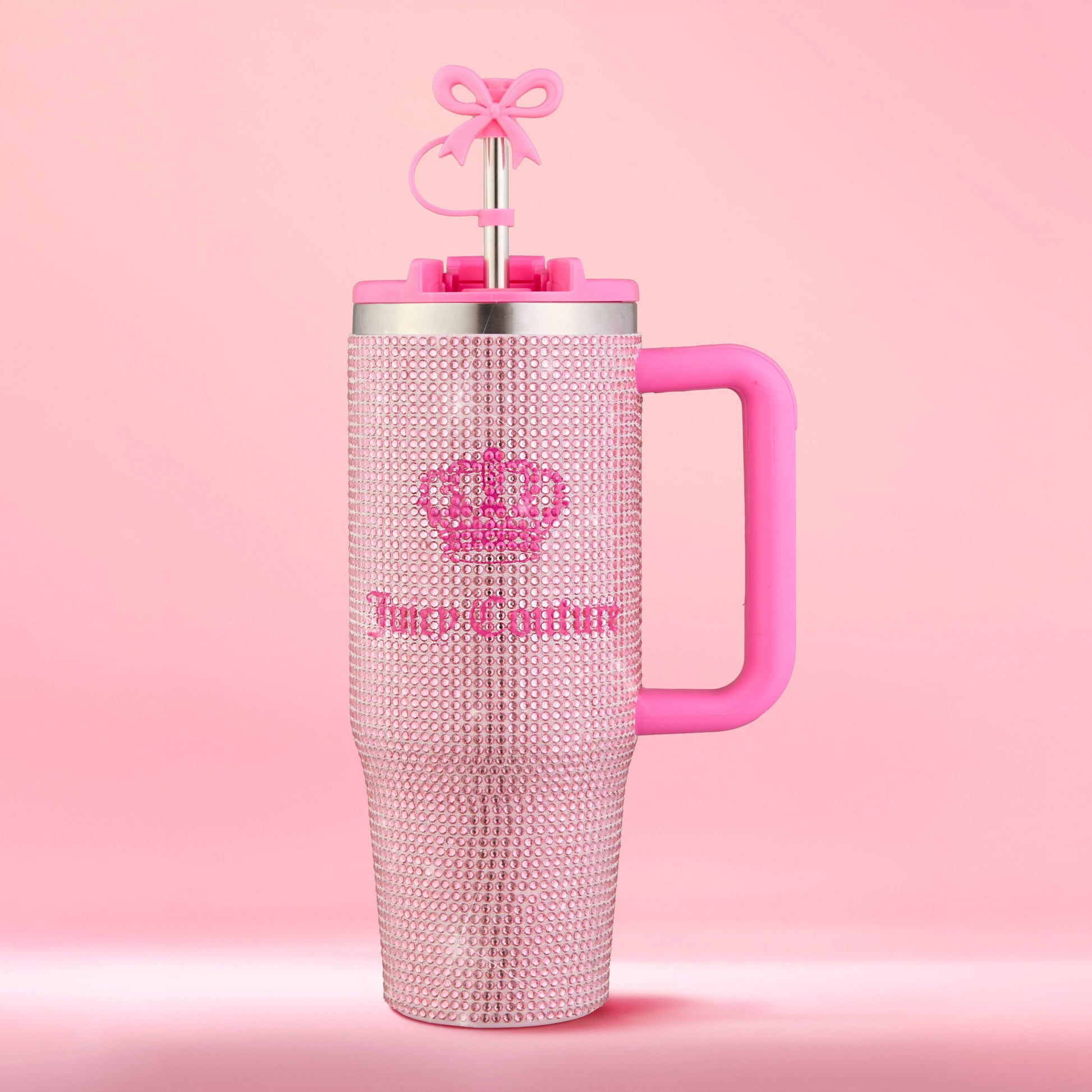 Introducing the Diamond Bling Stainless Steel Tumbler with Handle and Straw, 30 oz—a pink tumbler dazzling with rhinestones, adorned with a crown and the Juicy Couture logo. This glamorous cup shines brilliantly, featuring a sturdy pink handle and a straw topped by a cute pink bow, all set against a perfectly matching pink background.