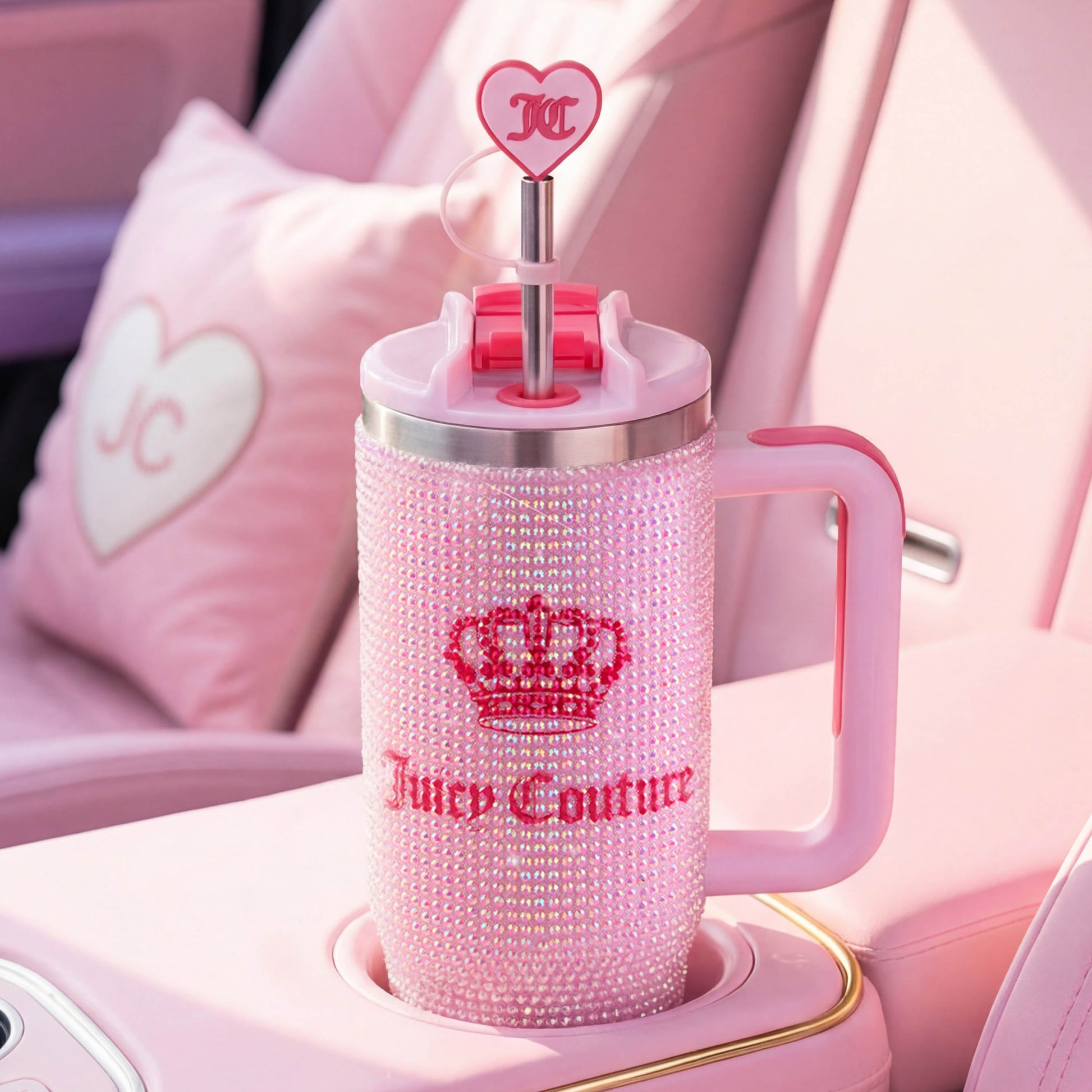 A Diamond Bling Stainless Steel Tumbler with Handle and Straw, 30 oz, sparkling with dazzling crystals, sits in a pink car cupholder. The tumbler comes in pink and features a crown logo along with a heart-shaped straw topper adorned with “JC.” A coordinating pink pillow rests on the car seat, completing the glamorous look.