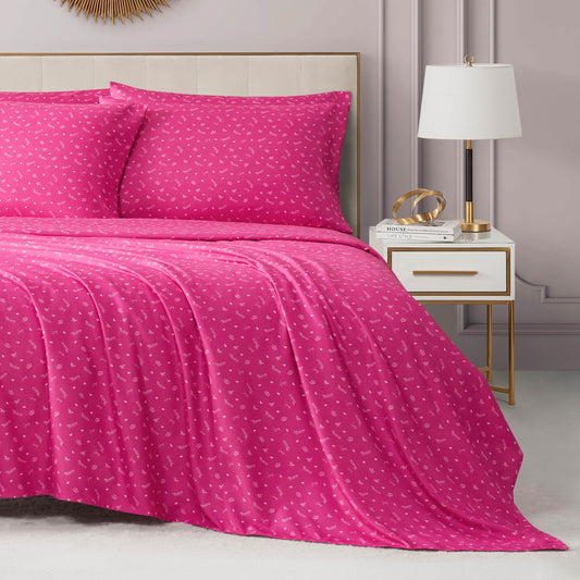 A stylish, light-toned bedroom features a neatly made bed dressed in the Microfiber Sheet Set With Extra Pillowcases in bright pink and a white heart pattern, next to a gold-accented nightstand with books, decor, and a modern lamp.