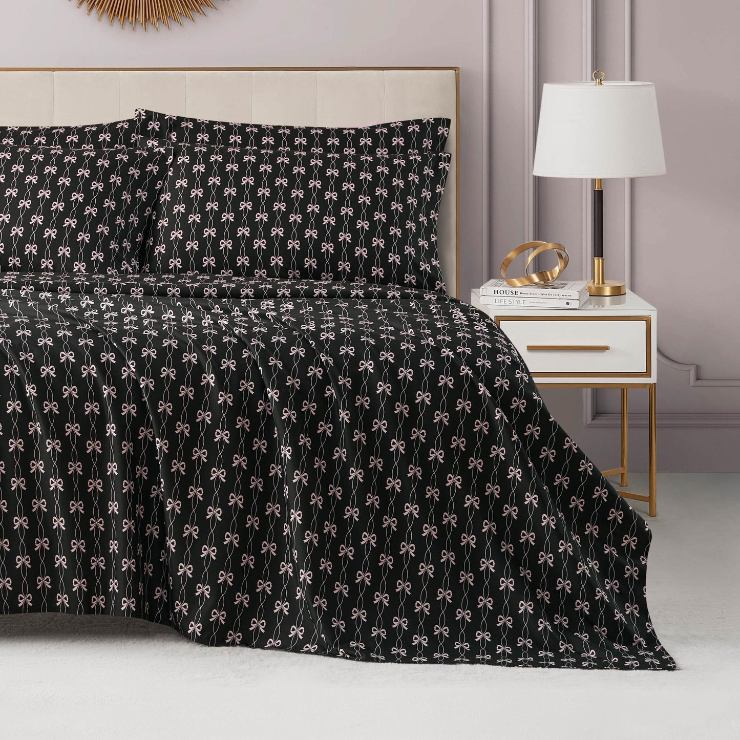 A modern bedroom features a neatly made bed adorned with a black microfiber sheet set with extra pillowcases displaying small white bows, complemented by luxury bedding, beside a white and gold nightstand with a lamp, books, and decor.