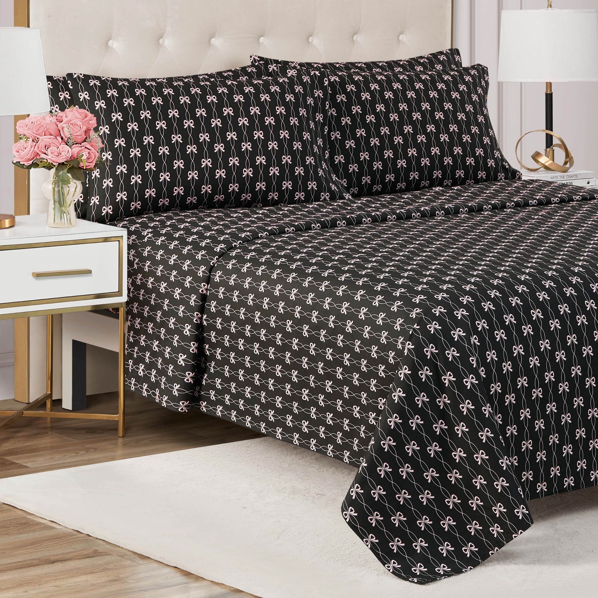 A neatly made bed with a black Microfiber Sheet Set With Extra Pillowcases featuring white bows sits against a tufted beige headboard. A white nightstand with gold trim holds pink flowers and books in this modern bedroom.