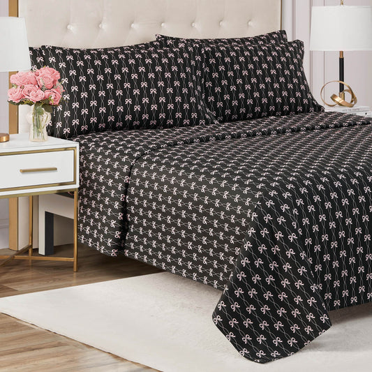 A neatly made bed with a black Microfiber Sheet Set With Extra Pillowcases featuring white bows sits against a tufted beige headboard. A white nightstand with gold trim holds pink flowers and books in this modern bedroom.