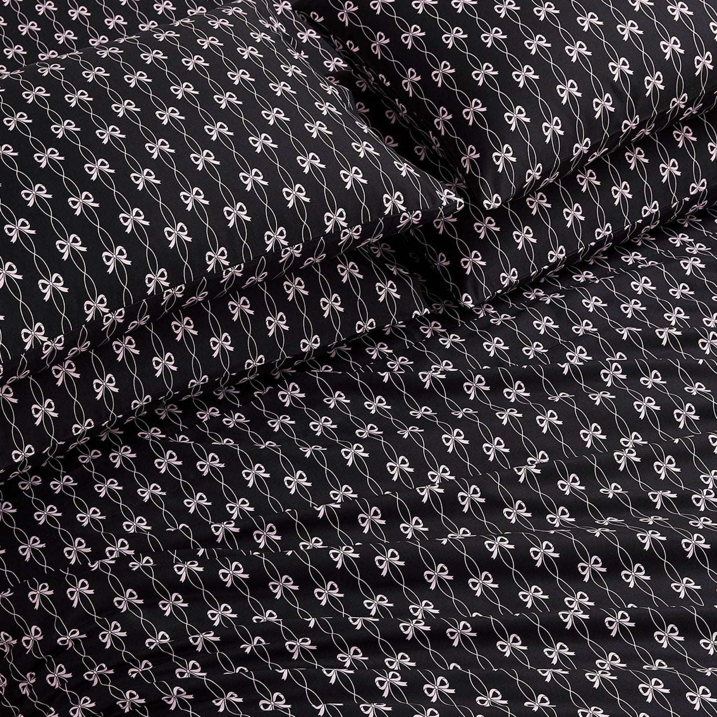The Microfiber Sheet Set With Extra Pillowcases features black luxury bedding with a repeating small white bow pattern, including a fitted sheet, flat sheet, and two pillowcases, all neatly arranged on a bed.