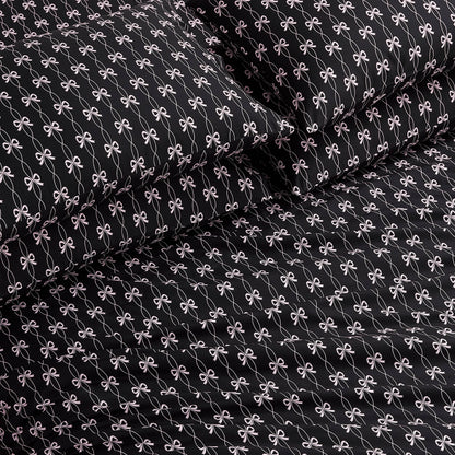 The Microfiber Sheet Set With Extra Pillowcases features black luxury bedding with a repeating small white bow pattern, including a fitted sheet, flat sheet, and two pillowcases, all neatly arranged on a bed.