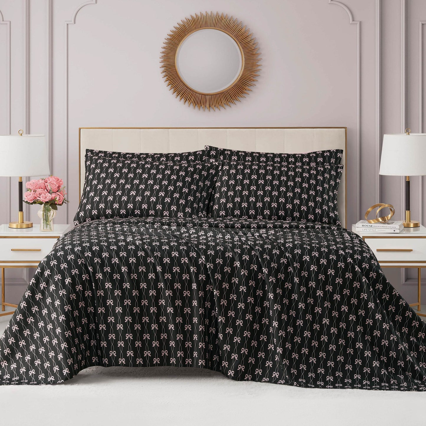 A neatly made bed with a black patterned comforter and matching pillows sits between two white nightstands, each with a lamp. A Microfiber Sheet Set With Extra Pillowcases adds comfort beneath a round sunburst mirror above the bed.