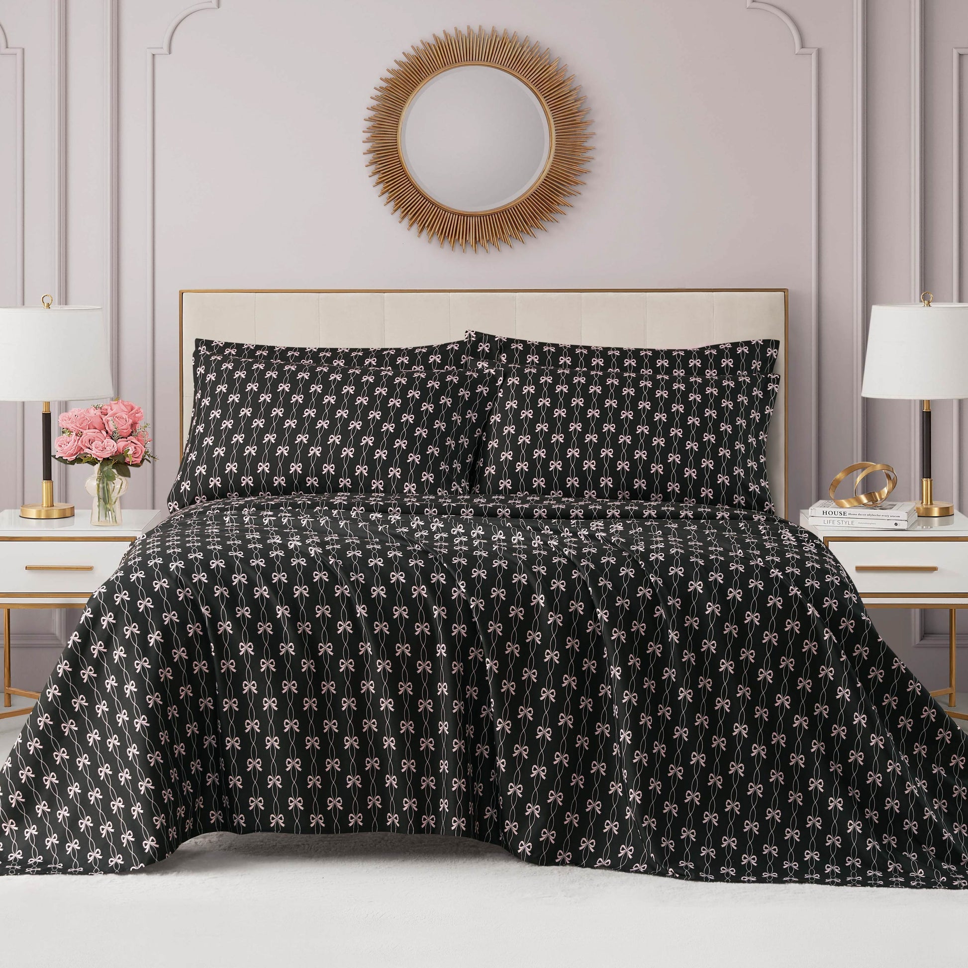 A neatly made bed with a black patterned comforter and matching pillows sits between two white nightstands, each with a lamp. A Microfiber Sheet Set With Extra Pillowcases adds comfort beneath a round sunburst mirror above the bed.