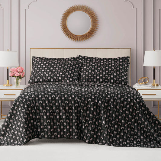 A neatly made bed with a black patterned comforter and matching pillows sits between two white nightstands, each with a lamp. A Microfiber Sheet Set With Extra Pillowcases adds comfort beneath a round sunburst mirror above the bed.