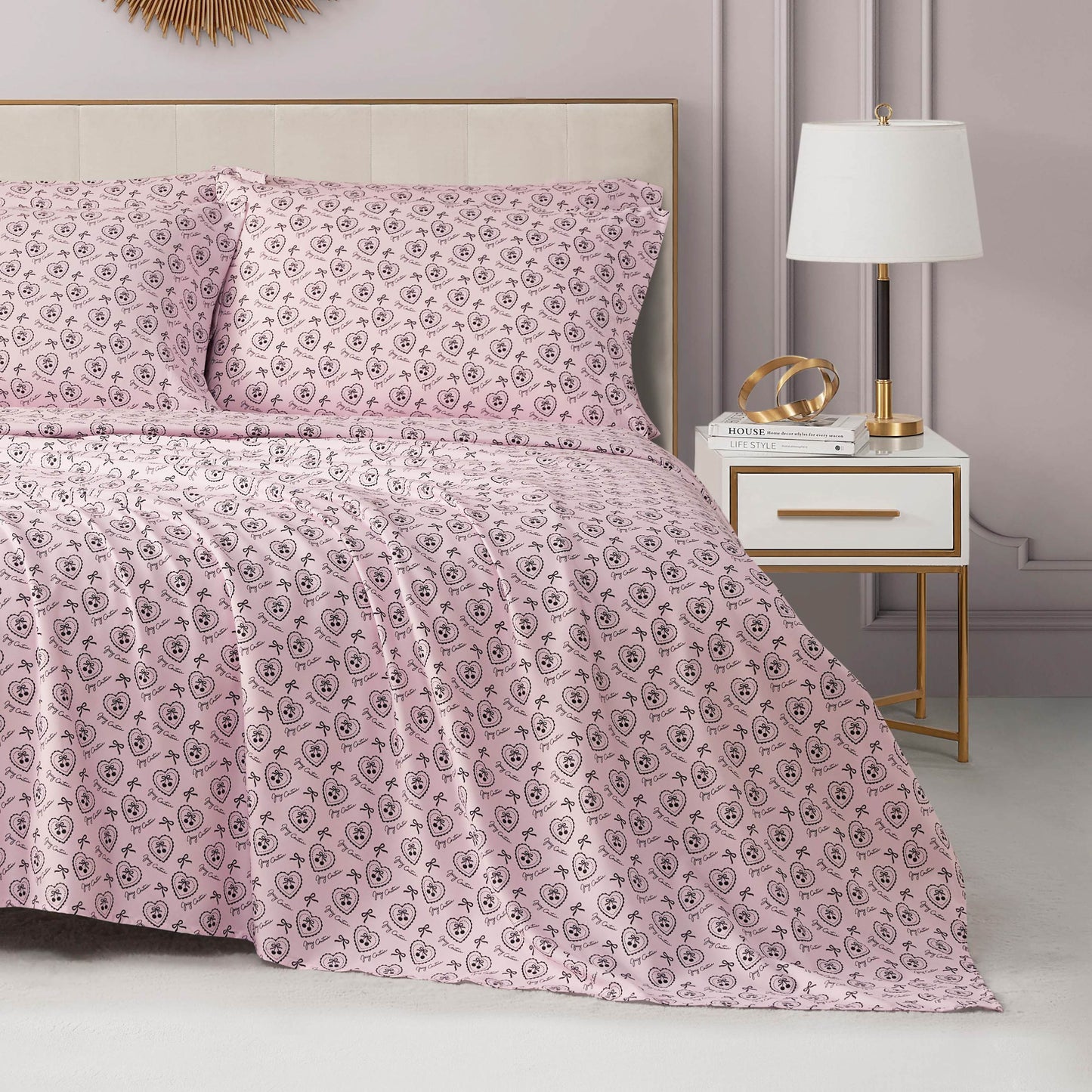 A neatly made bed with the Microfiber Sheet Set With Extra Pillowcases, featuring pink sheets and pillowcases with a small black floral pattern, is beside a white nightstand with gold accents. The room’s walls are light lavender.