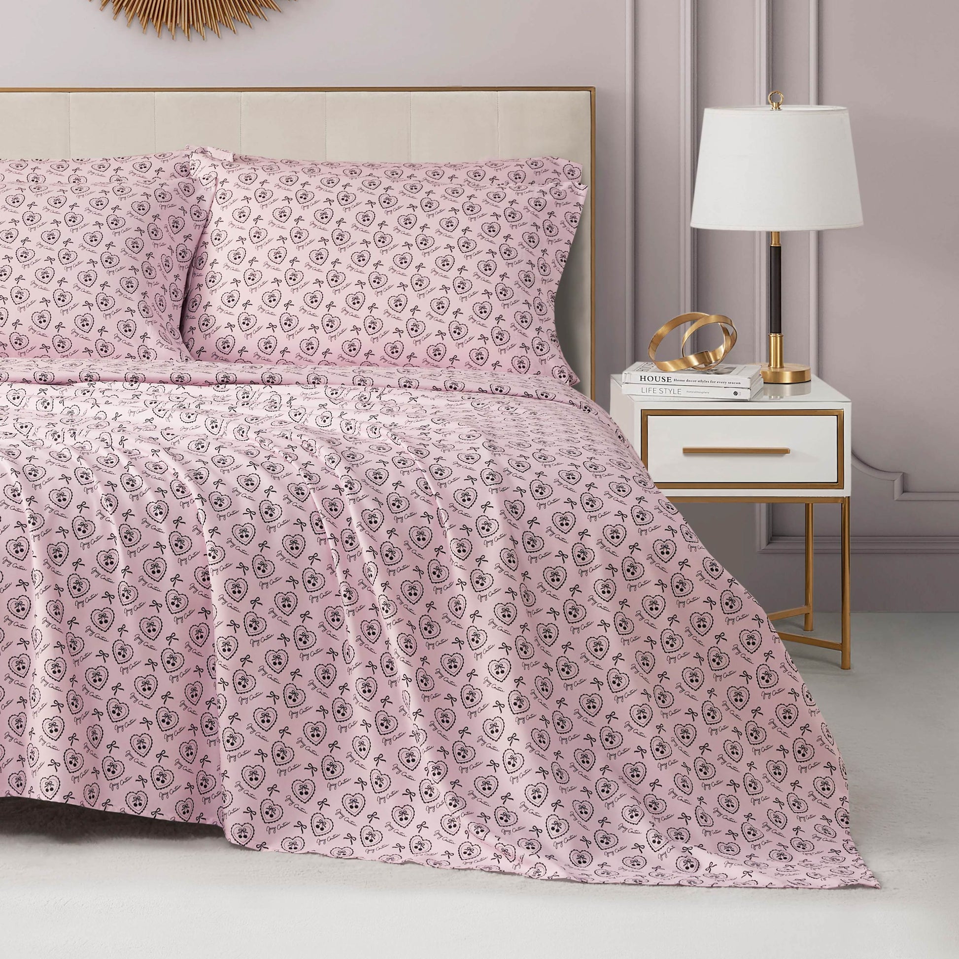A neatly made bed with the Microfiber Sheet Set With Extra Pillowcases, featuring pink sheets and pillowcases with a small black floral pattern, is beside a white nightstand with gold accents. The room’s walls are light lavender.