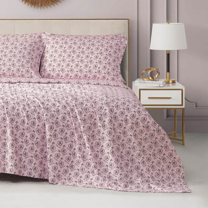 A neatly made bed with the Microfiber Sheet Set With Extra Pillowcases, featuring pink sheets and pillowcases with a small black floral pattern, is beside a white nightstand with gold accents. The room’s walls are light lavender.