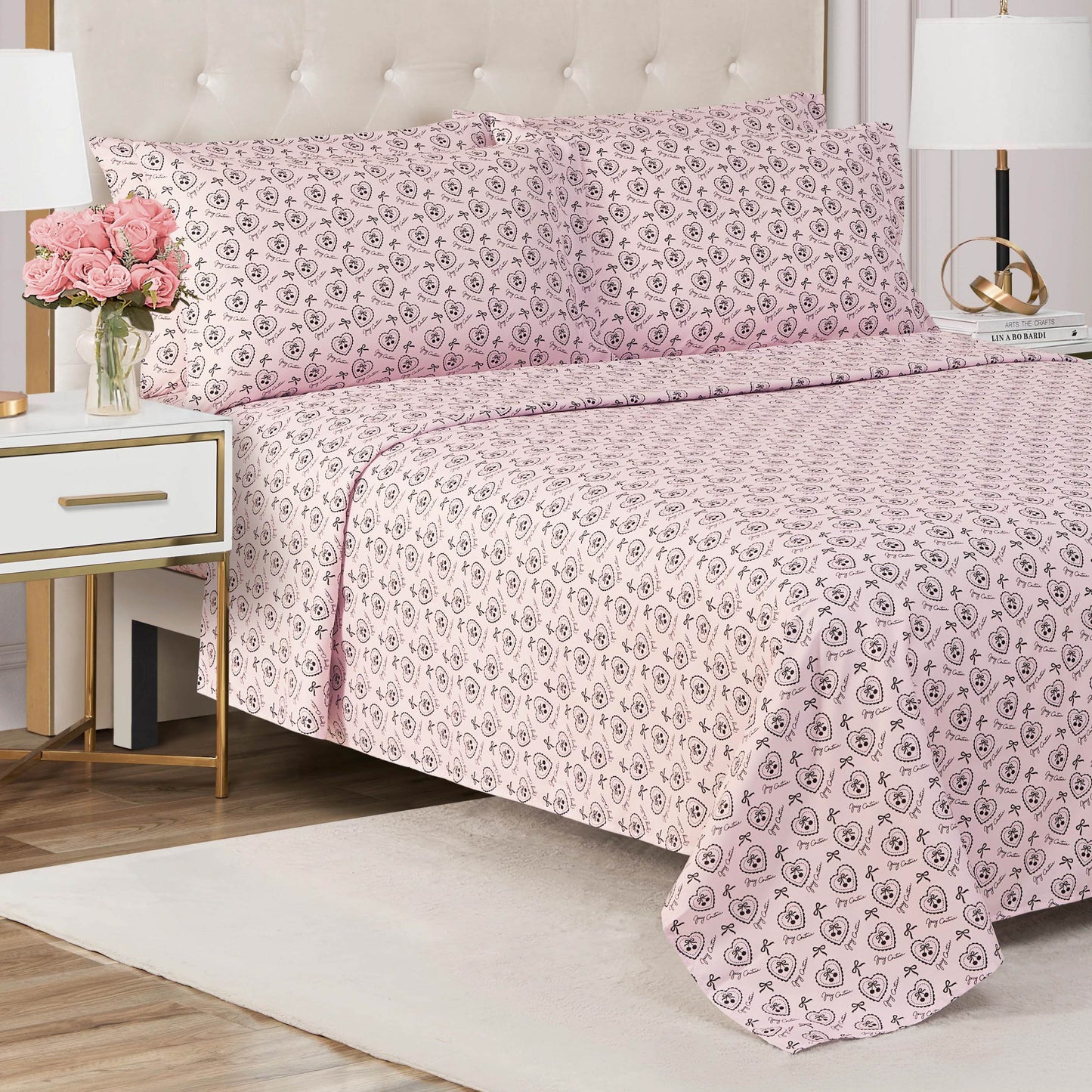 A neatly made bed with a Microfiber Sheet Set With Extra Pillowcases in pink floral patterns and matching pillowcases sits in a modern bedroom featuring a white nightstand, pink flowers, gold accents, and a white table lamp.