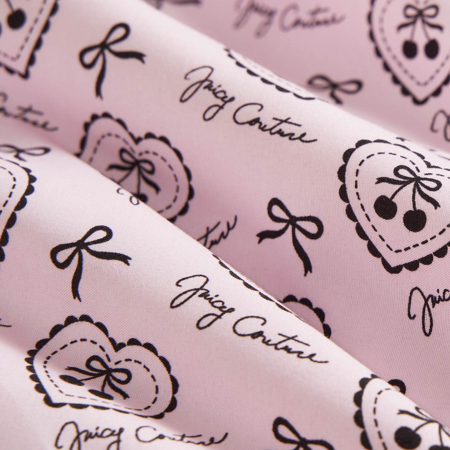 Close-up of the Microfiber Sheet Set With Extra Pillowcases in pink, featuring black heart, bow, and cherry illustrations plus Juicy Couture cursive. Soft and slightly folded, it adds a playful, chic touch to your bedroom.