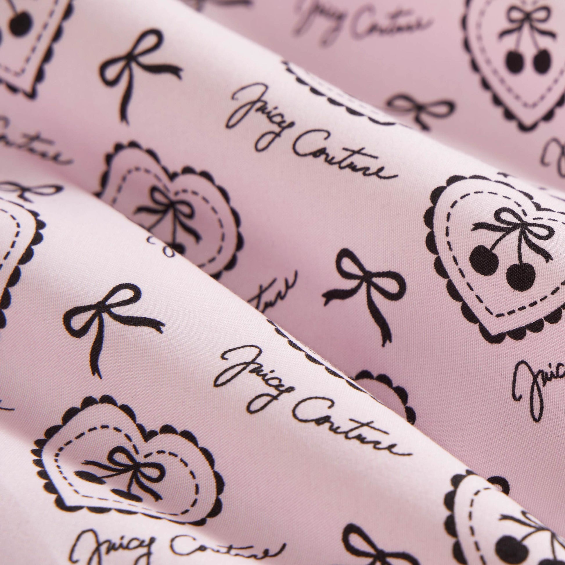 Close-up of the Microfiber Sheet Set With Extra Pillowcases in pink, featuring black heart, bow, and cherry illustrations plus Juicy Couture cursive. Soft and slightly folded, it adds a playful, chic touch to your bedroom.