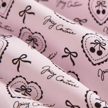 Close-up of the Microfiber Sheet Set With Extra Pillowcases in pink, featuring black heart, bow, and cherry illustrations plus Juicy Couture cursive. Soft and slightly folded, it adds a playful, chic touch to your bedroom.