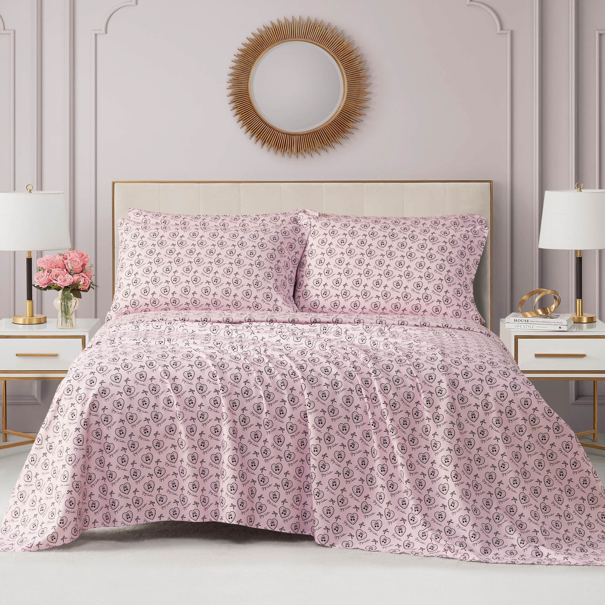A neatly made bed with a pink and white patterned coverlet and matching pillows sits against a light mauve wall, topped by a gold sunburst mirror. The Microfiber Sheet Set With Extra Pillowcases adds a luxurious, inviting touch.