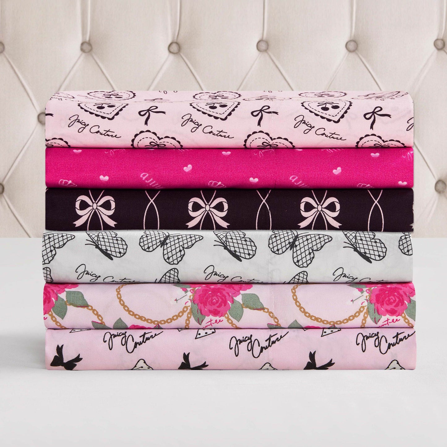 Five folded, patterned fabrics in pink, black, gray, and white—featuring bows, hearts, roses, teddy bears, and Juicy Couture text—showcase the Microfiber Sheet Set With Extra Pillowcases to elevate your luxury bedding against a tufted headboard.