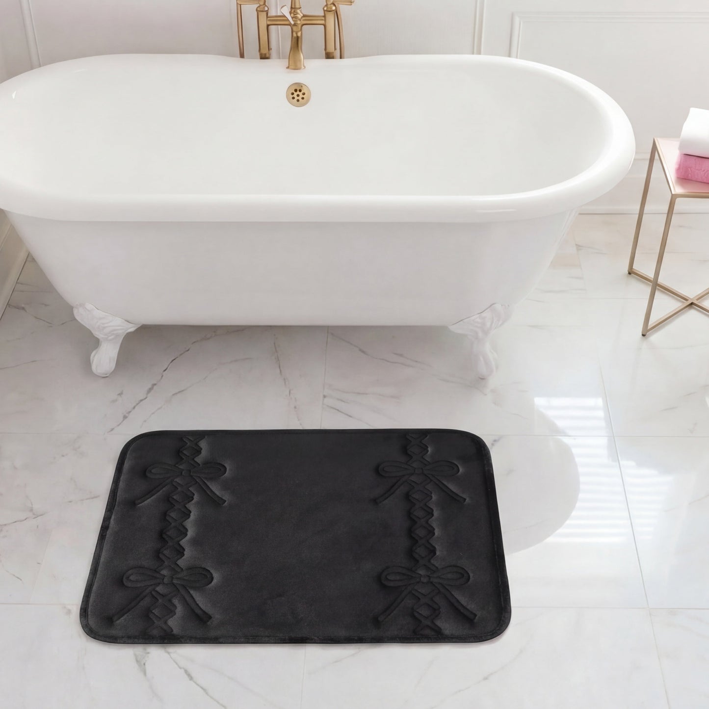 A white clawfoot bathtub with gold fixtures sits on marble flooring. In front of the tub is a black Quilted Memory Foam Bath Mat, 17" x 24". A side table holds a towel and a soap dispenser.