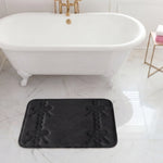 A white clawfoot bathtub with gold fixtures sits on marble flooring. In front of the tub is a black Quilted Memory Foam Bath Mat, 17" x 24". A side table holds a towel and a soap dispenser.