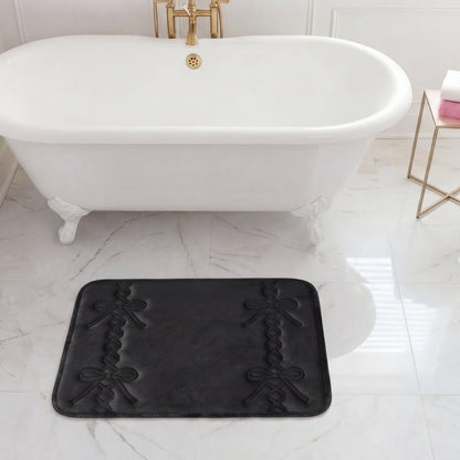 A white clawfoot bathtub with gold fixtures sits on marble flooring. In front of the tub is a black Quilted Memory Foam Bath Mat, 17" x 24". A side table holds a towel and a soap dispenser.