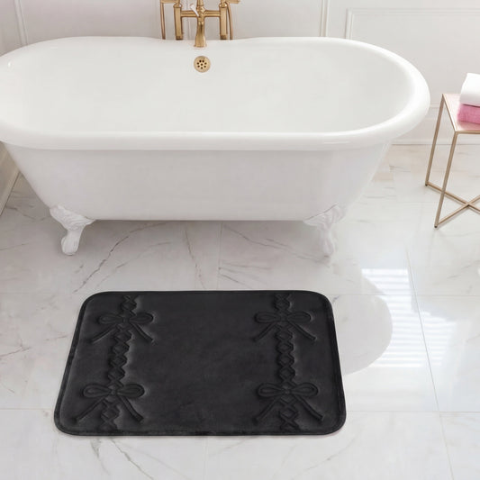 A white clawfoot bathtub with gold fixtures sits on marble flooring. In front of the tub is a black Quilted Memory Foam Bath Mat, 17" x 24". A side table holds a towel and a soap dispenser.