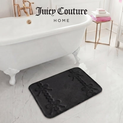 A luxurious bathroom features a white clawfoot tub, gold fixtures, and a 17" x 24" Quilted Memory Foam Bath Mat with black bows. Stacked white and pink towels add elegance, and "Juicy Couture Home" is displayed above the tub.