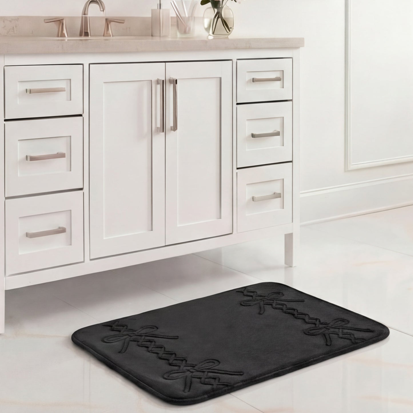 A black Quilted Memory Foam Bath Mat, 17" x 24", with a decorative pattern is placed on a white tiled floor in front of a white bathroom vanity with silver handles and a beige countertop.