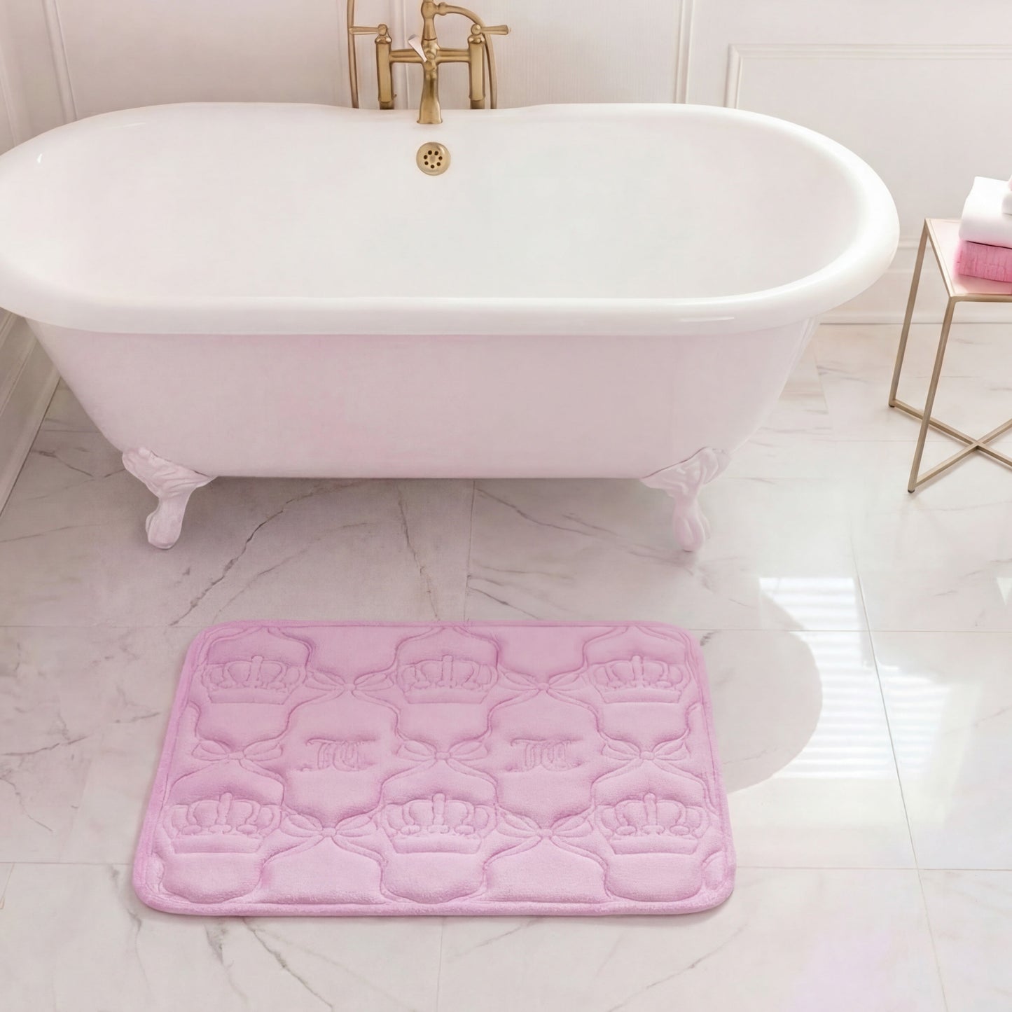 A white clawfoot bathtub with gold fixtures stands on a marble floor. In front of it is a pink Quilted Memory Foam Bath Mat, 17" x 24", with embossed crown designs. Towels are neatly stacked on a gold side table nearby.