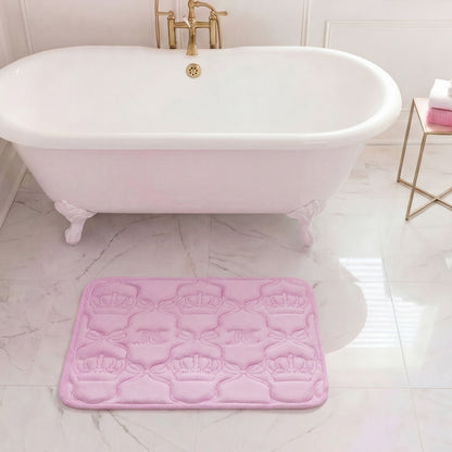 A white clawfoot bathtub with gold fixtures stands on a marble floor. In front of it is a pink Quilted Memory Foam Bath Mat, 17" x 24", with embossed crown designs. Towels are neatly stacked on a gold side table nearby.