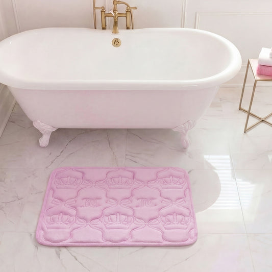 A white clawfoot bathtub with gold fixtures stands on a marble floor. In front of it is a pink Quilted Memory Foam Bath Mat, 17" x 24", with embossed crown designs. Towels are neatly stacked on a gold side table nearby.