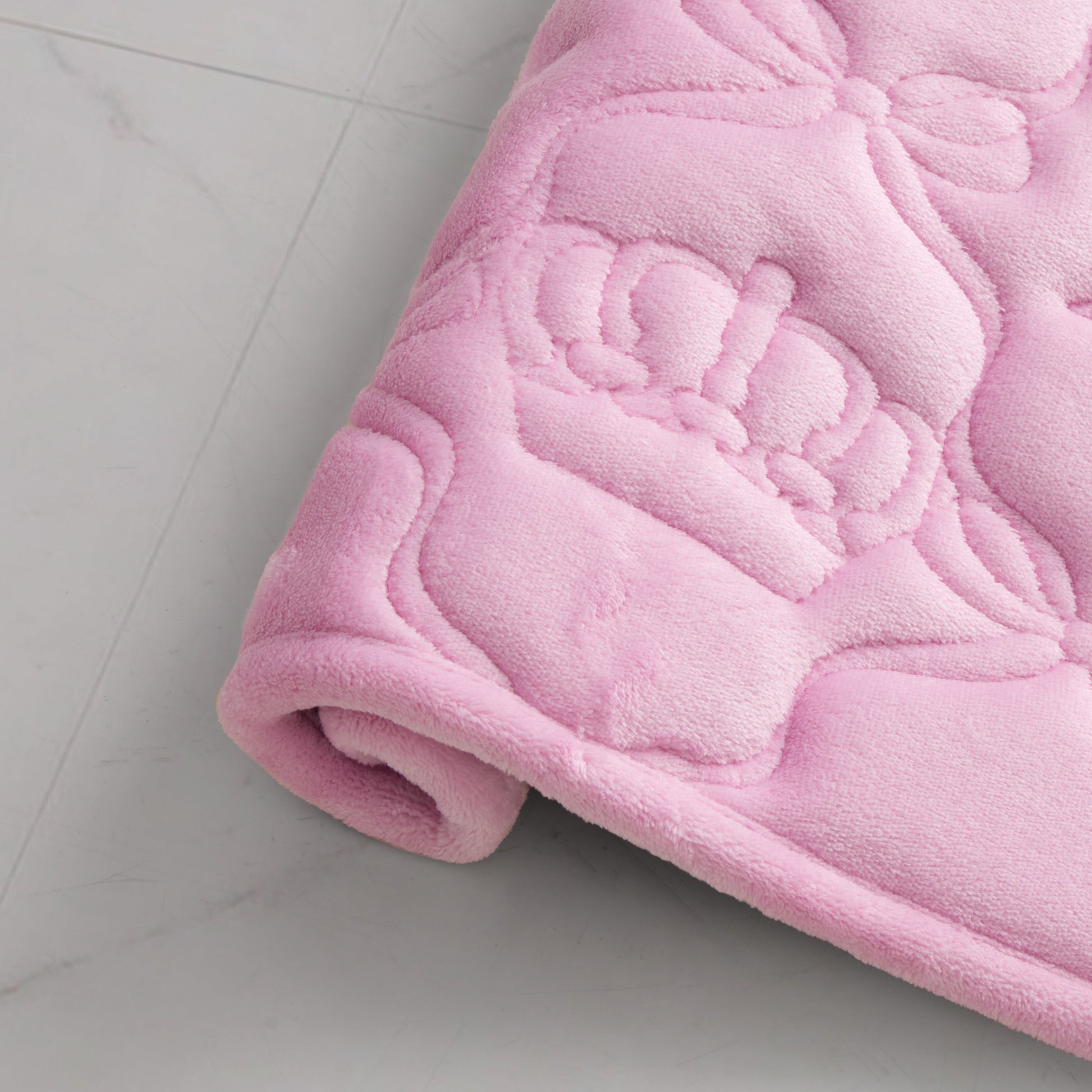 A soft, pink, rolled-up blanket with an embossed crown and bow design rests beside a Quilted Memory Foam Bath Mat, 17" x 24", on a light-colored tiled floor.