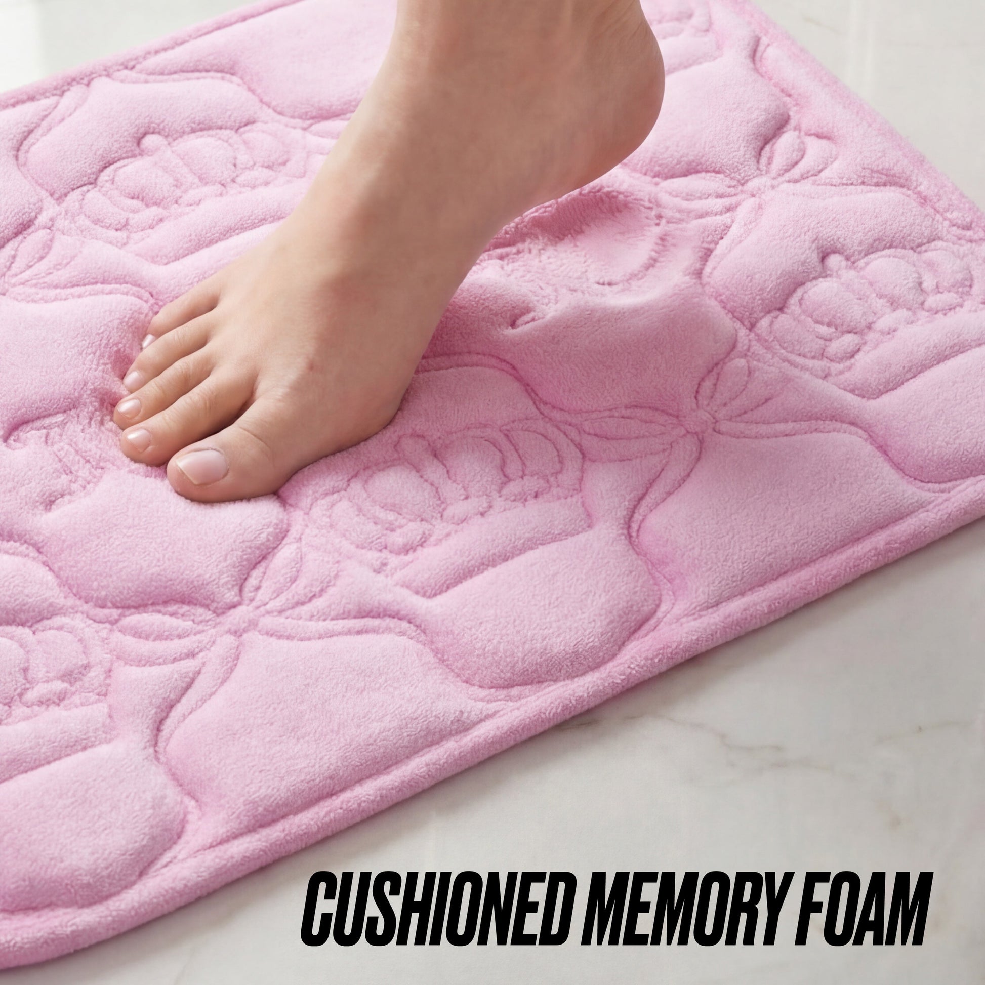 A bare foot steps on a soft, pink Quilted Memory Foam Bath Mat, 17" x 24", with flower patterns, placed on a light-colored floor. Bold text at the bottom reads: CUSHIONED MEMORY FOAM.