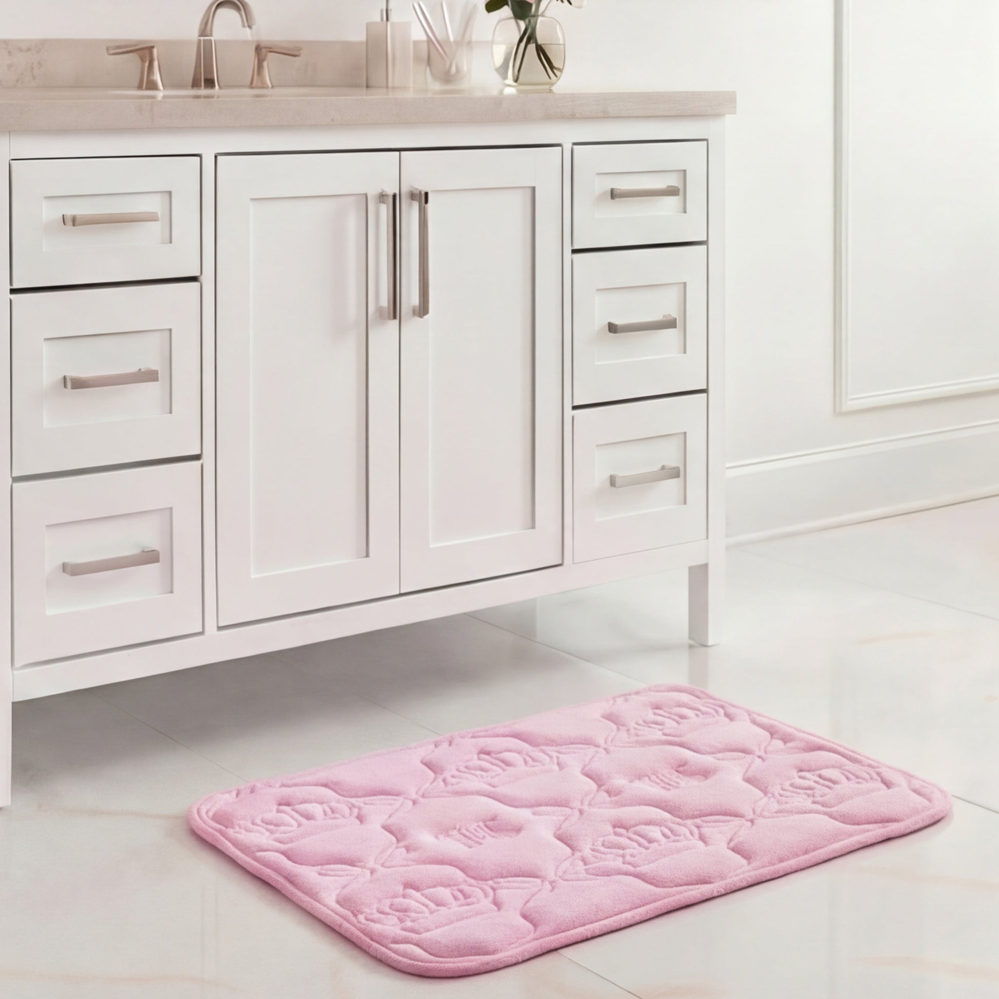 A Quilted Memory Foam Bath Mat, 17" x 24", in light pink with a floral pattern, is placed on a shiny white tile floor in front of a white bathroom vanity with silver handles and a gray countertop.