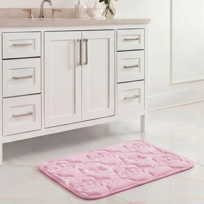 A Quilted Memory Foam Bath Mat, 17" x 24", in light pink with a floral pattern, is placed on a shiny white tile floor in front of a white bathroom vanity with silver handles and a gray countertop.