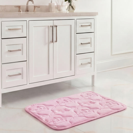 A Quilted Memory Foam Bath Mat, 17" x 24", in light pink with a floral pattern, is placed on a shiny white tile floor in front of a white bathroom vanity with silver handles and a gray countertop.