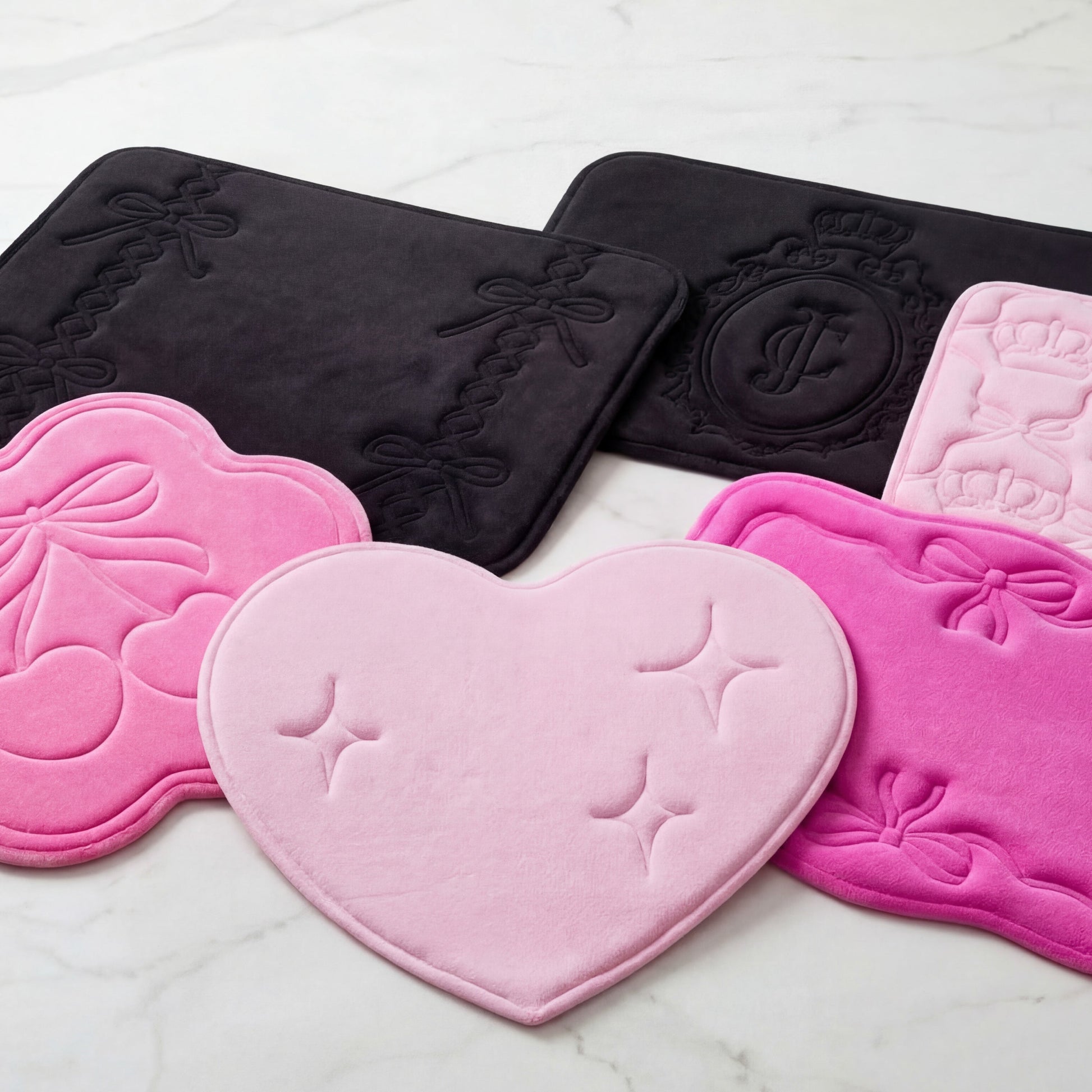 Five Quilted Memory Foam Bath Mats (17" x 24") in black, pink, and light pink feature embossed bows, crowns, cherries, and a heart. Each plush mat is shown on a white marble surface.
