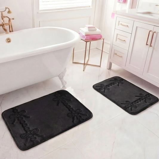 A bright bathroom features a white bathtub, gold fixtures, stacked towels on a stool, and the Quilted Memory Foam Bath Mat 2-Piece Set with bow patterns on a white tiled floor. White vanity cabinets are seen on the right.