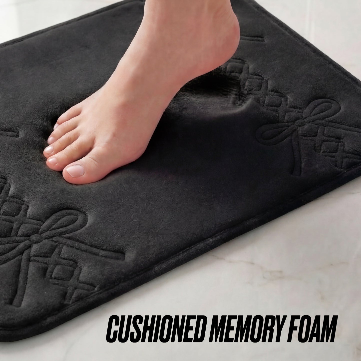 A bare foot steps on the black Quilted Memory Foam Bath Mat 2-Piece Set with bow patterns, showcasing its cushioned texture. The words CUSHIONED MEMORY FOAM appear at the bottom right, emphasizing this luxurious bathroom mat set.