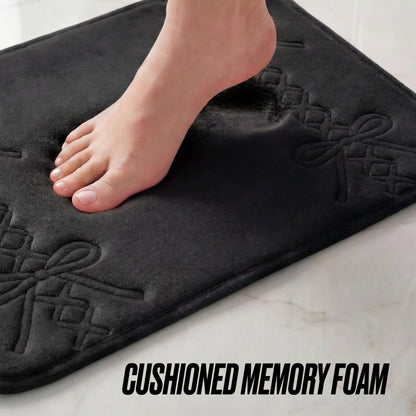 A bare foot steps on the black Quilted Memory Foam Bath Mat 2-Piece Set with bow patterns, showcasing its cushioned texture. The words CUSHIONED MEMORY FOAM appear at the bottom right, emphasizing this luxurious bathroom mat set.