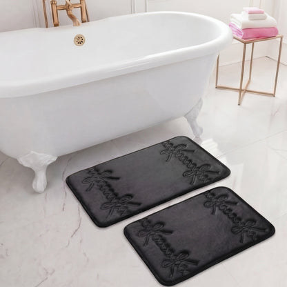 A white clawfoot bathtub stands on a glossy floor. In front is the Quilted Memory Foam Bath Mat 2-Piece Set with elegant cursive writing. A nearby side table displays folded white and pink towels.