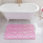 A white clawfoot bathtub sits in a marble-tiled bathroom with a plush Quilted Memory Foam Bath Mat (20" x 32") placed in front. A small gold side table nearby holds a white and pink bottle.