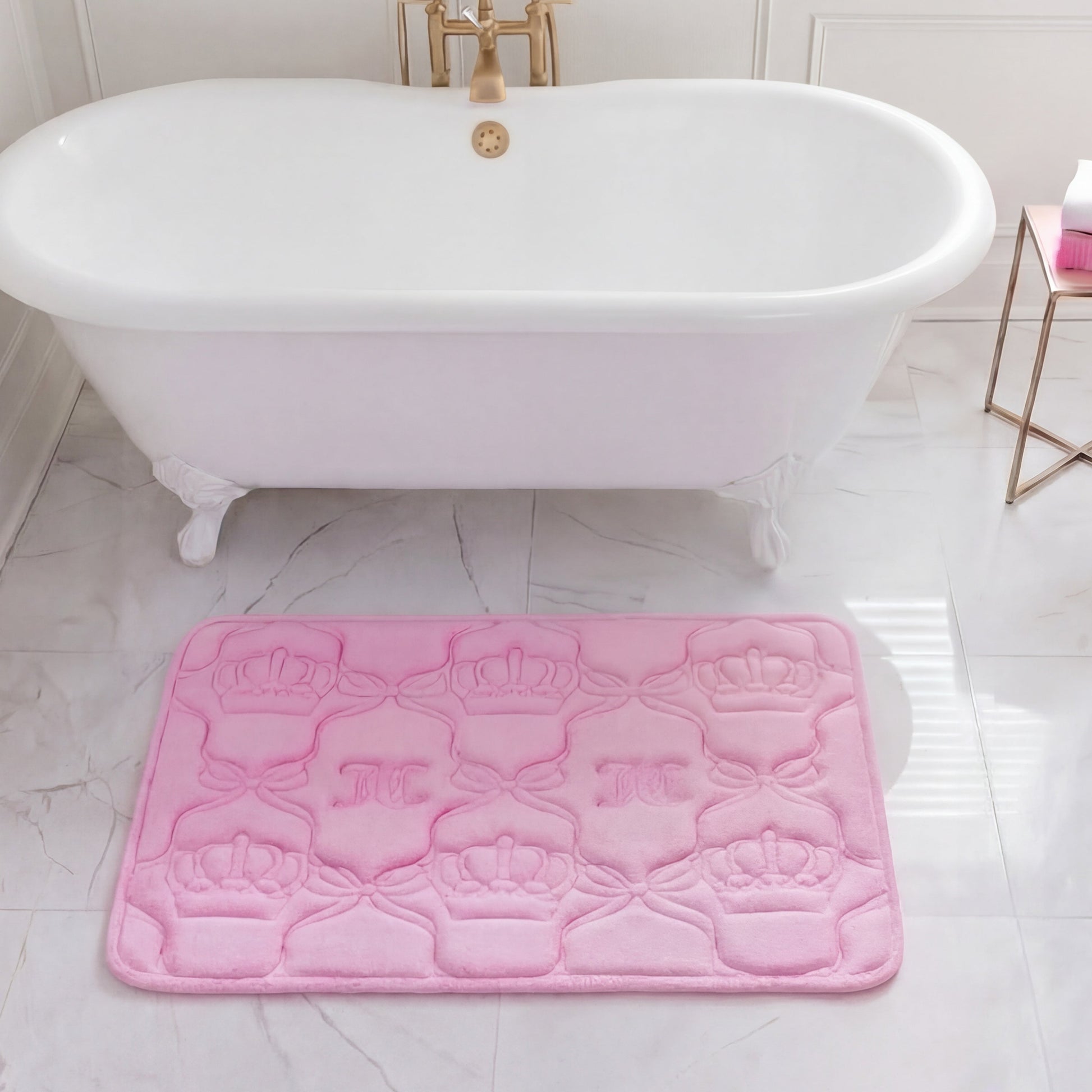A white clawfoot bathtub sits in a marble-tiled bathroom with a plush Quilted Memory Foam Bath Mat (20" x 32") placed in front. A small gold side table nearby holds a white and pink bottle.