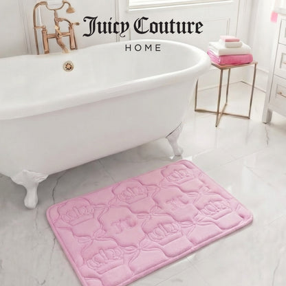 A luxurious white clawfoot tub with gold fixtures sits on a marble floor. A plush pink Juicy Couture Quilted Memory Foam Bath Mat (20" x 32") and neatly stacked pink and white towels add a stylish touch. "Juicy Couture HOME" is above the tub.