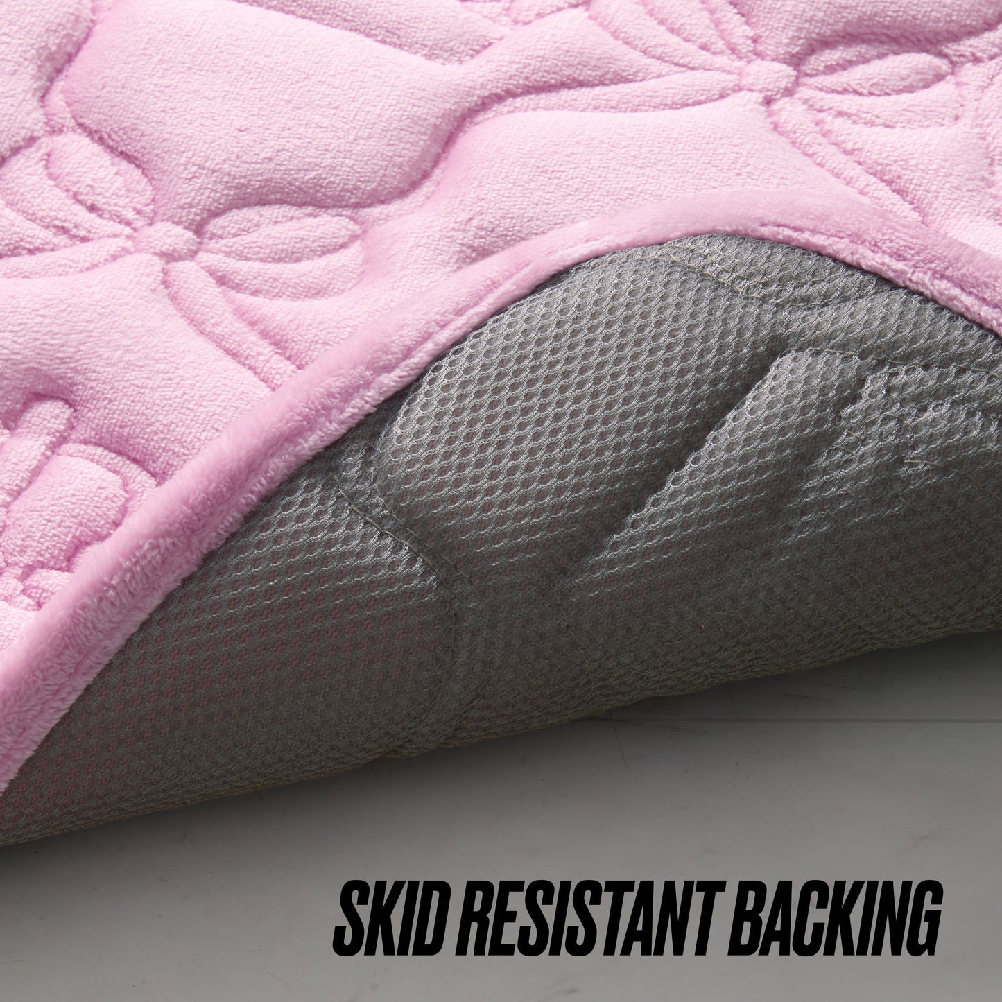 A close-up of the Quilted Memory Foam Bath Mat, 20" x 32", in pink, with the corner lifted to show its textured gray skid-resistant backing. The words SKID RESISTANT BACKING appear at the bottom right.