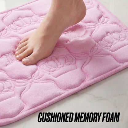 A bare foot steps onto a soft, pink Quilted Memory Foam Bath Mat, 20" x 32", with floral patterns, placed on a light-colored floor. Text at the bottom reads CUSHIONED MEMORY FOAM.