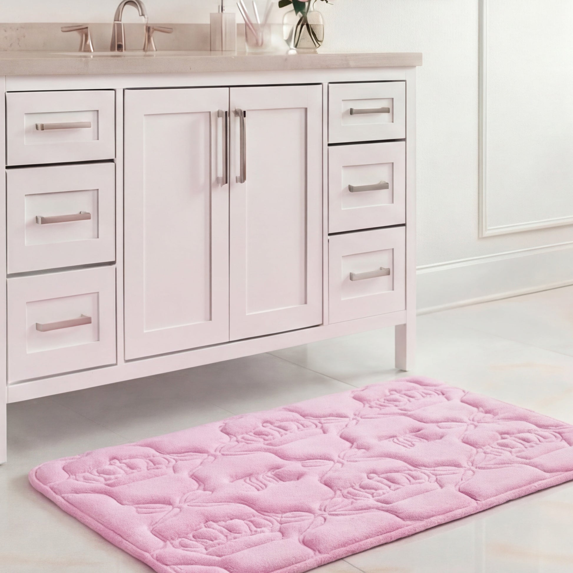 A Quilted Memory Foam Bath Mat, 20" x 32", in light pink with a floral pattern is placed on a glossy white tile floor in front of a white bathroom vanity with silver handles.